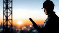 Industrial worker in hard hat using tablet at construction site during golden hour sunset with communication tower silhouette in background for project management.