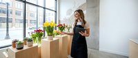 Young female florist in black apron arranging colorful spring flowers in modern shop interior with large windows and natural light for business.