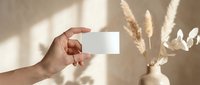 Hand holding blank white business card mockup with pampas grass decoration in soft natural lighting for branding presentation.