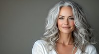 Confident mature woman with natural silver gray wavy hair and radiant smile wearing white sweater against neutral background, beauty and aging gracefully concept.