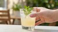 Person holding refreshing lemonade cocktail with fresh mint leaves outdoors on sunny day, perfect for summer beverage, drink recipe, and hospitality design projects.