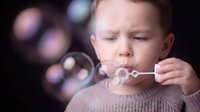 Young Caucasian boy blowing soap bubbles with colorful bokeh lights in background, childhood play and wonder concept for family lifestyle content.
