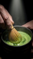 Person with dark skin tone whisking matcha green tea powder in traditional ceramic bowl using bamboo whisk, creating frothy ceremonial beverage for Japanese tea ceremony preparation.