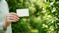 Woman in light green shirt holding blank white business card outdoors with blurred green foliage background, suitable for branding and corporate marketing materials.