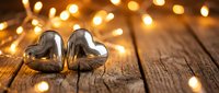 Two silver heart-shaped ornaments on rustic wooden surface with warm golden bokeh lights creating romantic festive atmosphere for holidays and celebrations.