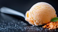 Creamy orange sherbet scoop on dark background with mint leaf garnish and crushed ice crystals, showcasing smooth texture and refreshing frozen dessert appeal.
