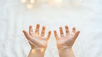 Hands holding glowing fairy lights string on soft white background, warm bokeh glow, magic and celebration concept.