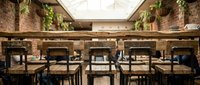 Modern restaurant interior with brick walls, wooden chairs, and hanging plants under skylight ceiling.