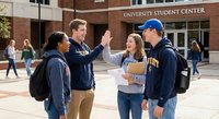 Diverse college students celebrating with high five outside university student center building on sunny campus day with backpacks and books for education success.