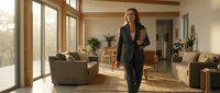 Young woman with long dark hair, South Asian ethnicity, wearing a black blazer and dark pants, walking confidently through a modern living room with wooden beams, large windows, and contemporary furniture during daytime.
