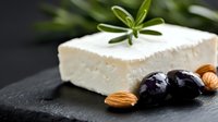 Fresh feta cheese block garnished with black olives, almonds and rosemary sprig on dark stone background, close up view with selective focus.