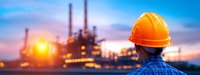 Industrial engineer in protective helmet observing oil refinery plant at sunset, panoramic view with illuminated petrochemical facility and glowing sky.
