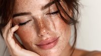 Young woman with freckles touching face in natural light showing healthy glowing skin and natural beauty for skincare and wellness concepts.
