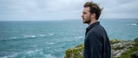 Bearded man with curly brown hair, Caucasian, wearing dark jacket, standing on rocky coastal cliff overlooking turbulent ocean waters on overcast day.