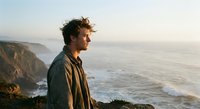 Young man with curly hair standing on coastal cliff overlooking ocean waves at sunset, contemplating peaceful seascape for wellness and mindfulness content.