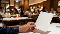 Close-up of mature male hand holding blank white card at formal dinner event with guests and elegant venue in soft-focus background for business, greeting, or announcement purposes.