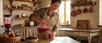 Woman in apron pouring fresh berry smoothie into glass in rustic kitchen with wooden shelves and hanging cookware for healthy lifestyle and food preparation content.
