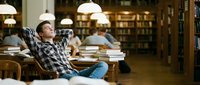 Young male student relaxing with hands behind head at library desk surrounded by stacks of books, taking a study break in academic environment.