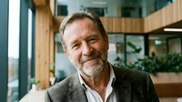 Middle-aged Caucasian man with gray beard wearing olive blazer and white shirt, smiling in modern office lobby with natural light and plants, suitable for corporate, business, and professional contexts.