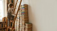 Young boy climbing wooden ladder to reach tall stack of books in cozy home library setting for educational and learning concepts.