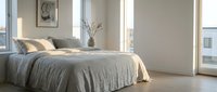 Serene minimalist bedroom with unmade bed, neutral linens, modern artwork, potted plant, and natural light from large windows.