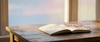 Open book with eyeglasses on wooden table by window with sunset sky view, suitable for education, reading, and lifestyle content.