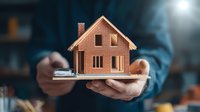 Hands holding miniature brick house model on white surface against dark background, illustrating real estate, property investment and home ownership concepts.