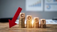 Business cost reduction concept with wooden blocks spelling COST on decreasing coin stacks and red arrow pointing down in modern office setting.