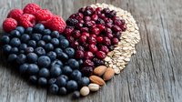 Heart-shaped arrangement of fresh berries, nuts and oats on rustic wooden background, featuring blueberries, raspberries, cranberries and almonds for healthy eating concept.