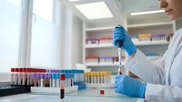 Female laboratory technician in white coat using pipette for blood sample analysis in modern medical research facility with test tubes and equipment.
