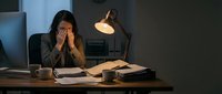 Exhausted businesswoman working late at night in dark office, holding head in hands showing stress and fatigue from overtime work and deadline pressure.