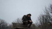 Young man in military uniform sitting alone on wooden bench in park during overcast day reflecting solitude and contemplation outdoors.