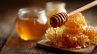 Golden honey dripping from wooden dipper onto honeycomb on rustic wooden board with jar of fresh honey in background.