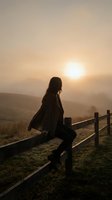 Woman with long dark hair sitting on wooden fence at sunrise over misty mountain valley, wearing dark jacket. Solitude and contemplation concept.