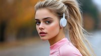 Young blonde woman wearing white headphones and pink shirt listening to music outdoors with autumn background for lifestyle and technology concepts.