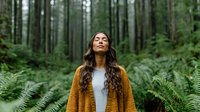 Young woman with long dark hair in mustard cardigan standing among ferns in peaceful forest, eyes closed in meditation or mindfulness moment.