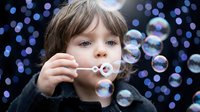 Young child with brown hair blowing soap bubbles against dark background with colorful bokeh lights creating magical atmosphere for childhood concepts.