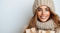 Young Caucasian woman in beige knit hat and scarf holding warm beverage cup, smiling at camera against light background for winter lifestyle content.