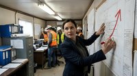Businesswoman in dark blazer pointing at upward trending arrow on whiteboard in construction site office with workers in safety vests and hard hats in background, representing business growth and project success.