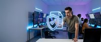 Young male cybersecurity professional in modern tech workspace with neon shield icon, multiple monitors, and team members working on data protection and network security solutions.