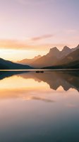 Serene mountain lake at sunrise with golden light reflecting on calm water, dramatic peaks in background, small boat on water for travel and nature concepts.