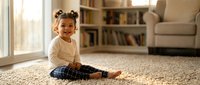 Happy mixed-race toddler girl with pigtails sitting on carpet in bright living room, smiling at camera. Child development and family lifestyle concept.