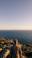 Man standing on rocky cliff overlooking calm ocean at sunset, wearing casual jacket, contemplating seascape with golden hour lighting and peaceful horizon view.