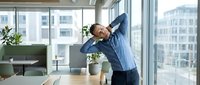 Young African American businessman stretching at modern office desk during work break. Professional male employee doing neck and shoulder exercises in corporate workspace.
