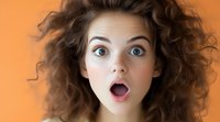 Young Caucasian woman with curly brown hair expressing extreme surprise with wide open mouth and eyes against orange background, emotional portrait closeup.