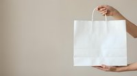 Hands holding blank white paper shopping bag against neutral background for retail branding and packaging mockup designs.