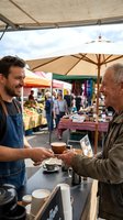 Caucasian male barista in denim apron serving coffee to senior Caucasian male customer at outdoor market cafe under beige umbrella, bustling farmers market background with vendors and shoppers.