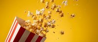Popcorn kernels flying out of red and white striped container against bright yellow background for entertainment and snack food concepts.