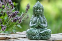 Green jade Buddha statue in meditation pose surrounded by purple wildflowers on wooden surface, creating peaceful zen garden atmosphere for spiritual and wellness settings.