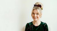 Young woman with blonde hair styled in an updo with decorative sunglasses on top, wearing dark green velvet top, smiling at camera against white background.